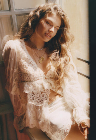 A woman with long wavy light brown hair wearing a sheer cream lace lingerie set with a matching long-sleeve robe, posed in warm sunlight near a window.